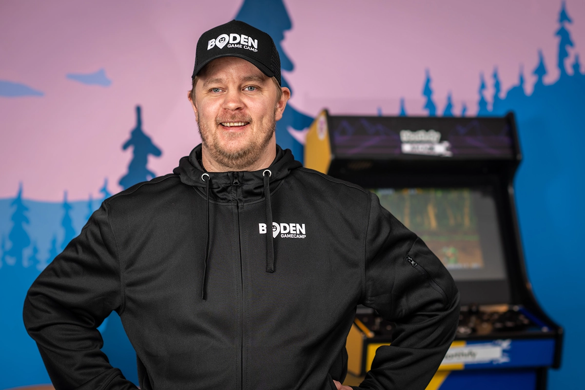 Man i svart hoodie och keps med logotypen 'Boden Game Camp' står leende framför en arkadmaskin och en vägg med färgglad skogssiluett i blått och rosa.