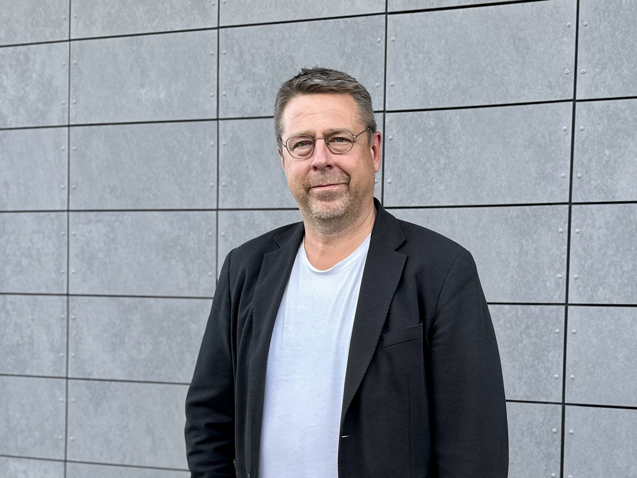 Man i glasögon, vit t-shirt och svart kavaj står framför en grå fasad med stora rektangulära paneler. Han ler lätt och tittar in i kameran.