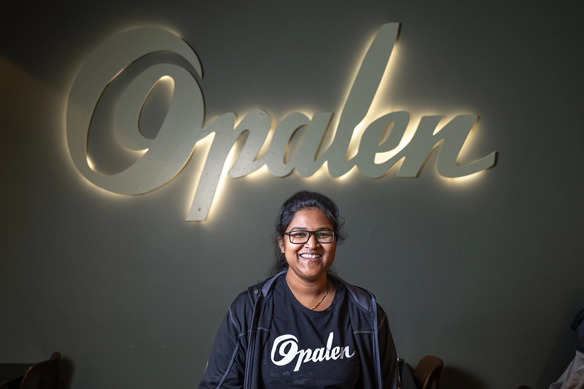 Kvinna i svart Opalen-tröja står leende framför en mörk vägg med den upplysta logotypen 'Opalen' i bakgrunden.