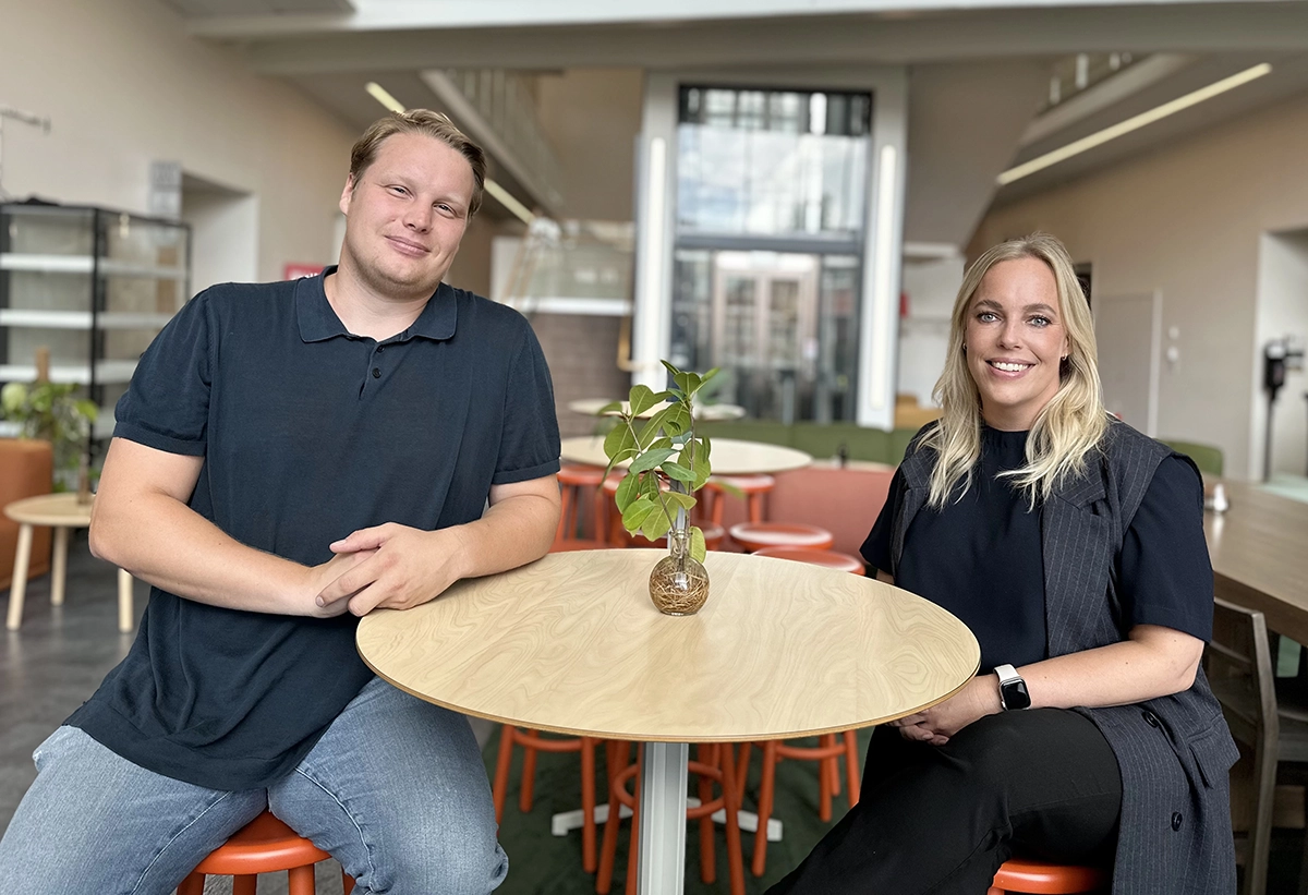 En man och en kvinna sitter vid ett runt bord i en ljus och modern lokal. Båda ler mot kameran. På bordet står en liten glasvas med gröna blad.