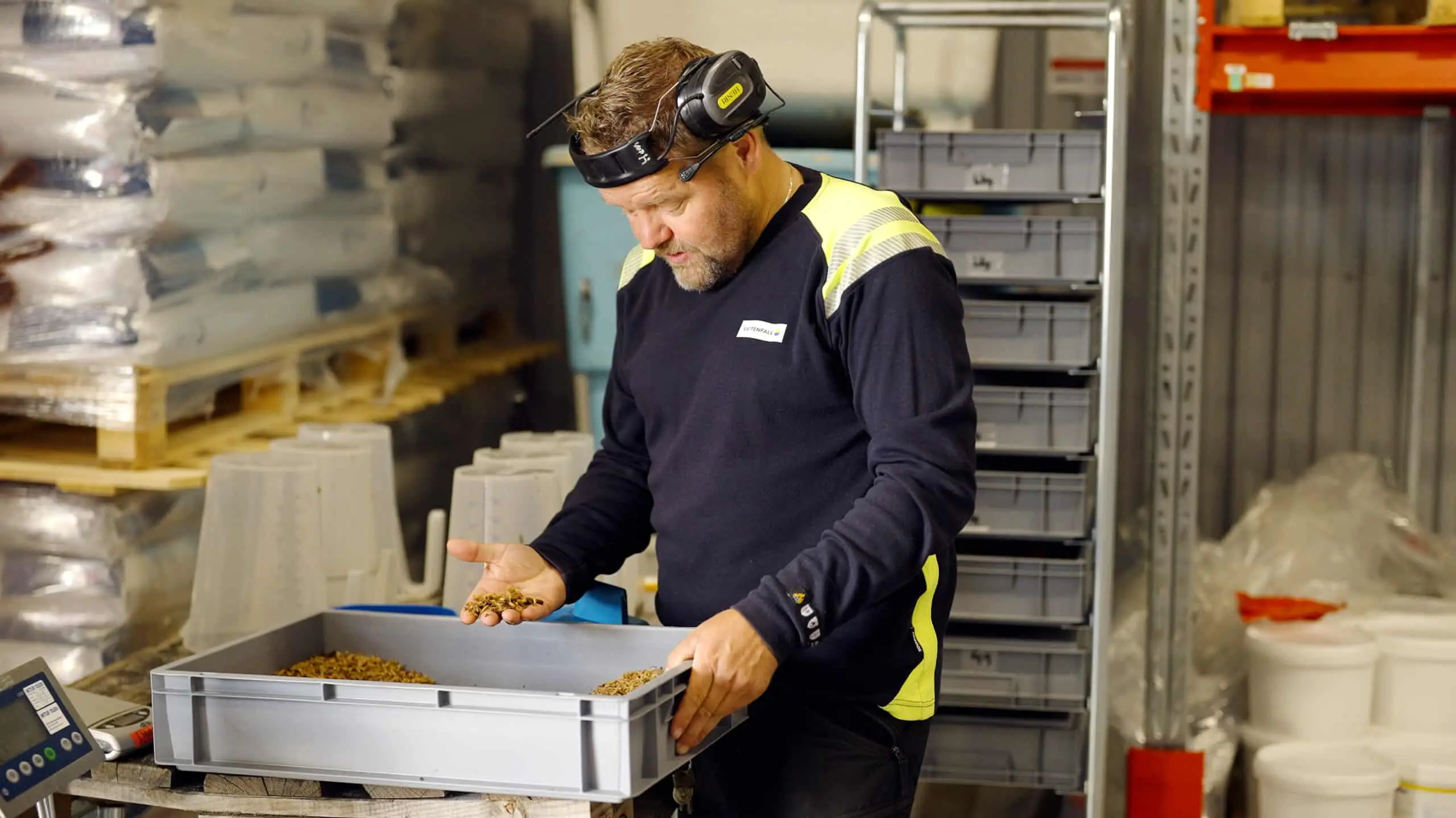Man i arbetskläder och hörselkåpor granskar fiskfoder i en grå plastlåda inne i ett lagerutrymme med hyllor och säckar i bakgrunden