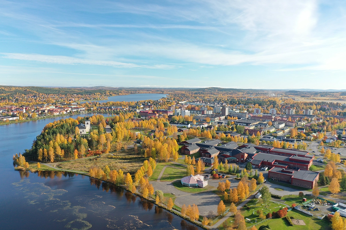 Flygfoto över Boden i höstskrud med färgsprakande träd, vattendrag och stadens byggnader. I förgrunden syns kyrkan på en udde och Bodens gymnasieområde med röda byggnader.