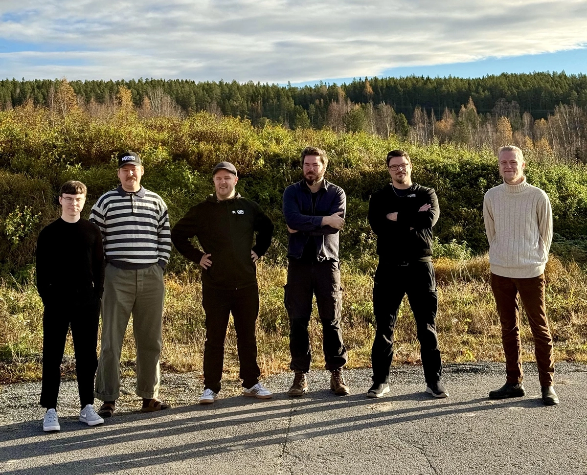 Sex män står uppradade utomhus i höstsolen med en skogsklädd sluttning i bakgrunden. De tittar mot kameran och ser avslappnade ut."