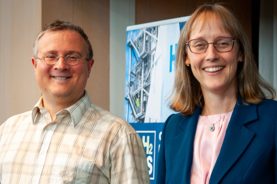 Leende man och kvinna med glasögon står framför en rollup med text och bild relaterad till vätgas. Mannen bär rutig skjorta och kvinnan en blå kavaj över rosa blus."