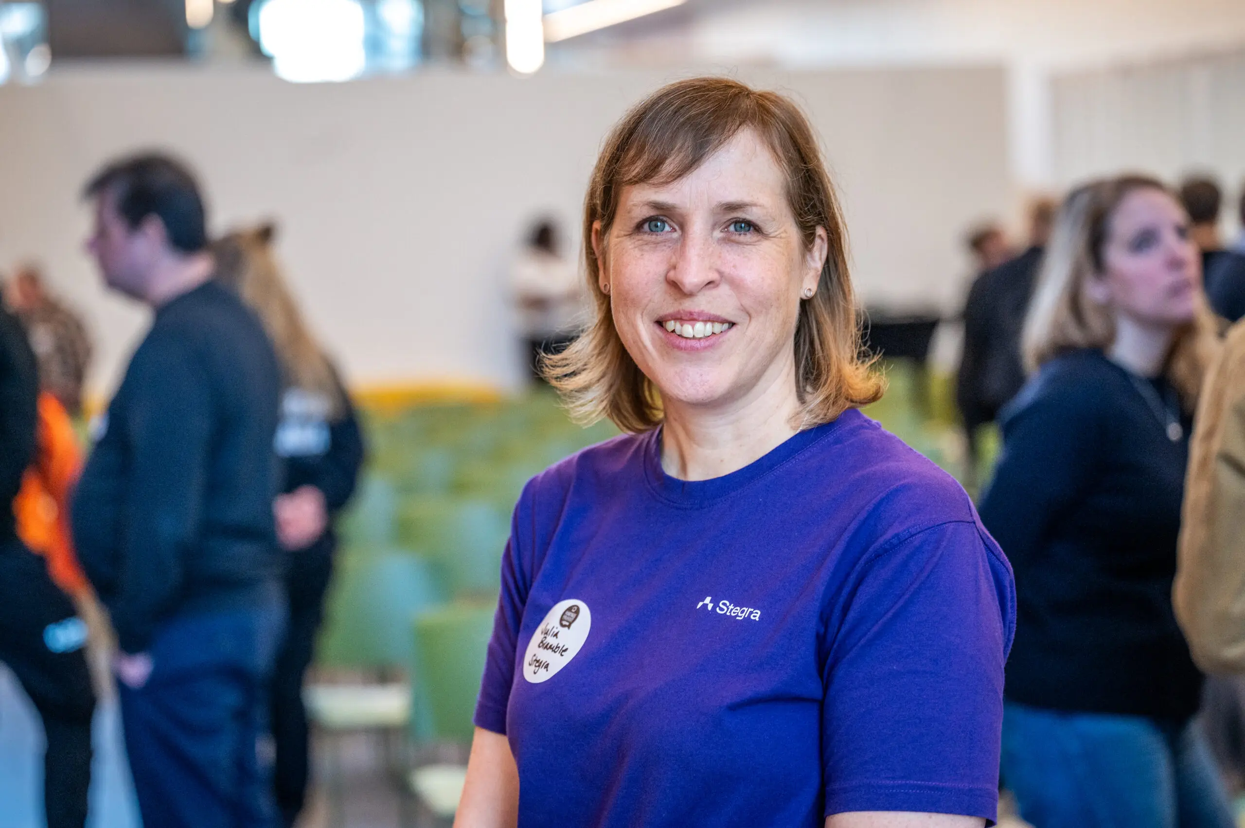 Leende kvinna i lila Stegra-tröja står i en ljus konferenslokal med gröna stolar och andra deltagare i bakgrunden.