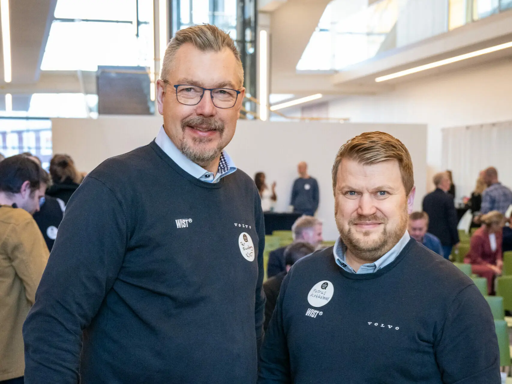 Två män i matchande mörkblå tröjor med logotyperna 'Wist' och 'Volvo' står leende i en ljus konferenslokal med andra deltagare i bakgrunden.