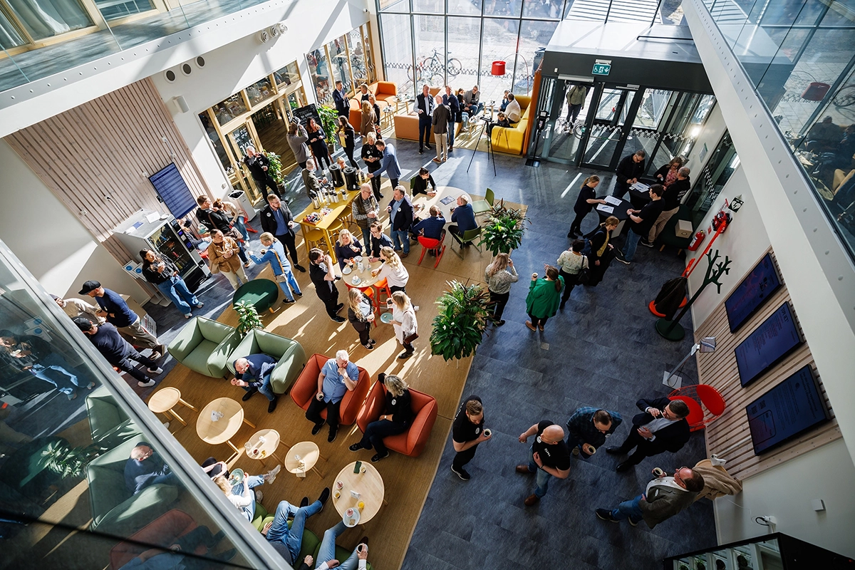 Överblicksbild från ovan av ett välbesökt event i en ljus och modern entréhall. Människor minglar, sitter i soffor och står i kö vid kaffestationer.